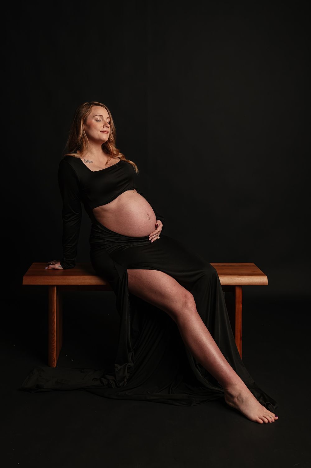 A dramatic maternity photography series in black dress against dark backdrop on wooden bench shows feminine silhouettes.