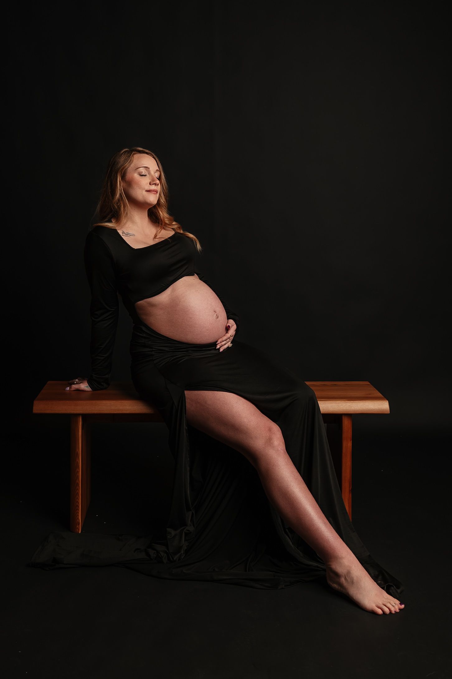 A dramatic maternity photography series in black dress against dark backdrop on wooden bench shows feminine silhouettes.