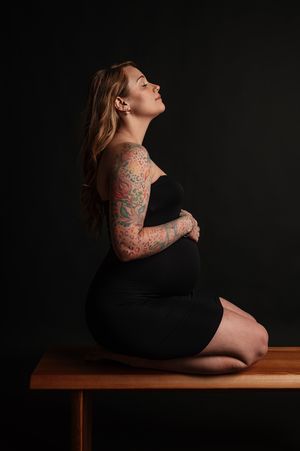 A person in a black dress poses artistically on a wooden surface against a dark background showing tattoos.