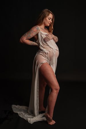 Woman in flowing beige dress poses against dark background showing artistic tattoo sleeve in dramatic lighting.