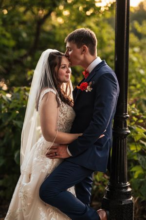Romantic wedding couple embraces in garden setting with evening sunlight filtering through trees.