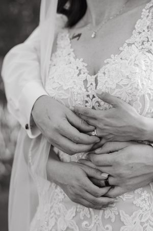 Black and white close-up of hands with wedding rings against delicate white lace wedding dress details.