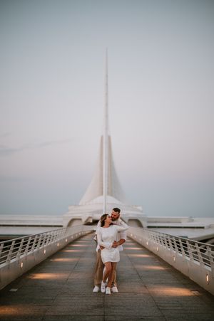 A romantic couple in white outfits poses together on the sleek Milwaukee Art Museum bridge at sunset.