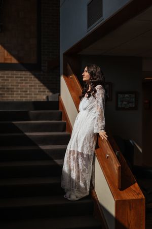 A person in a flowing white lace dress leans against a wooden railing on a dark staircase in dramatic lighting.