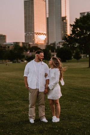 A couple in casual white outfits walks together on grassy field with urban buildings in background during sunset.