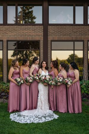 Bridal party in mauve bridesmaid dresses and white wedding gown pose together on grass outside brick building.