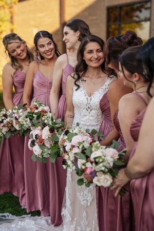 A bridal party wearing dusty rose bridesmaid dresses gathers outdoors holding pink and white floral bouquets.