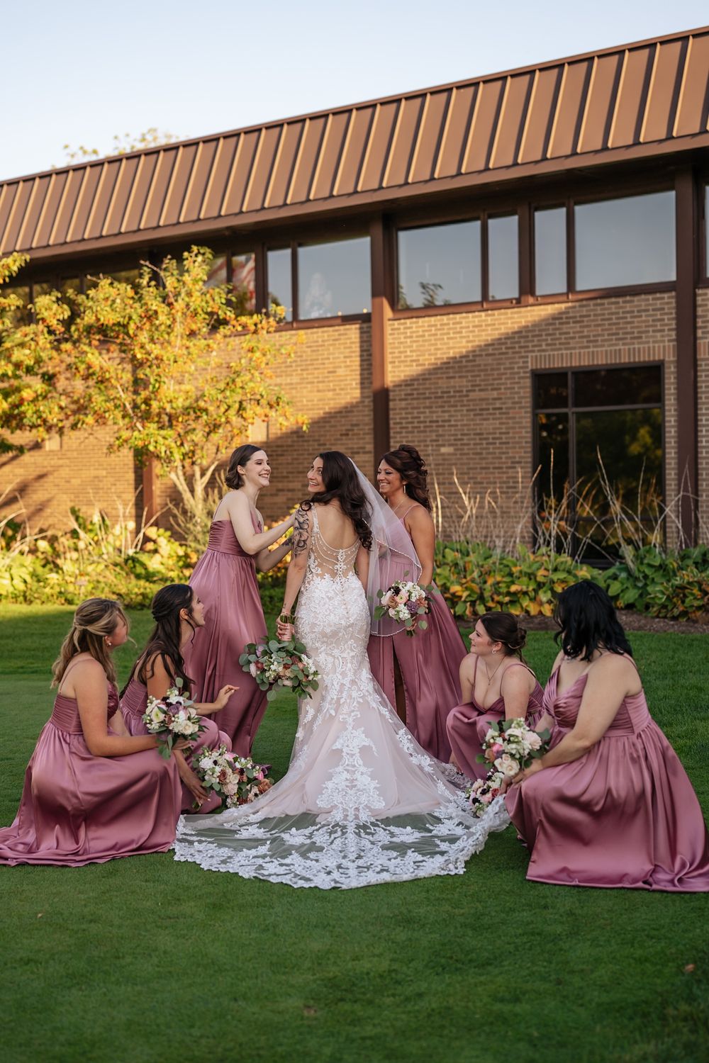 A wedding party in mauve bridesmaid dresses and white bridal gown sits together on lush green grass at sunset.