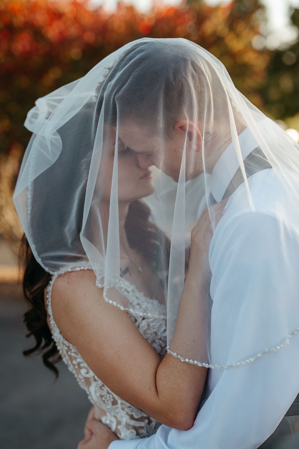 Romantic wedding couple shares intimate kiss under flowing white veil during golden autumn sunset.