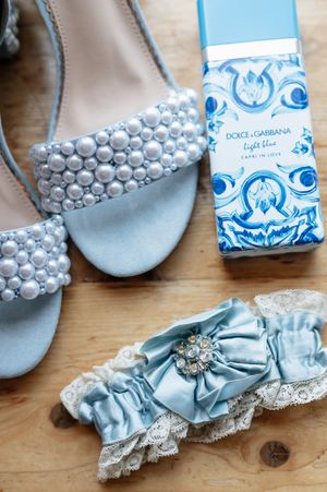 Light blue bridal accessories including pearl-embellished shoes, garter, and Dolce & Gabbana perfume on wooden surface.