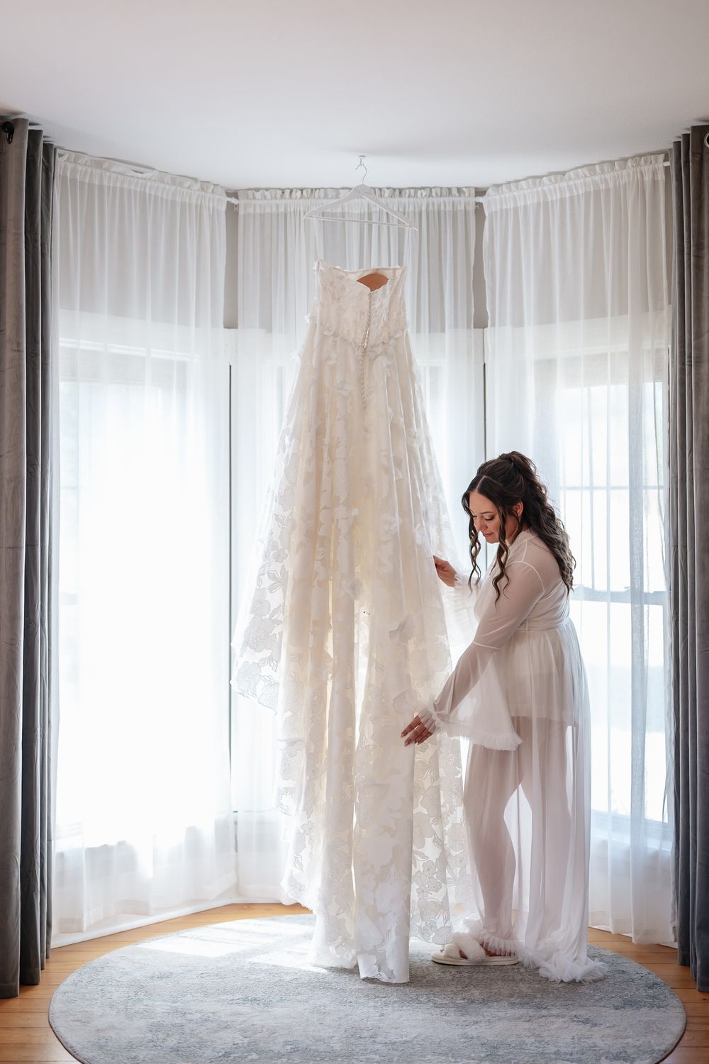 A delicate lace wedding dress hangs by a window as someone in a white robe admires it in soft natural light.