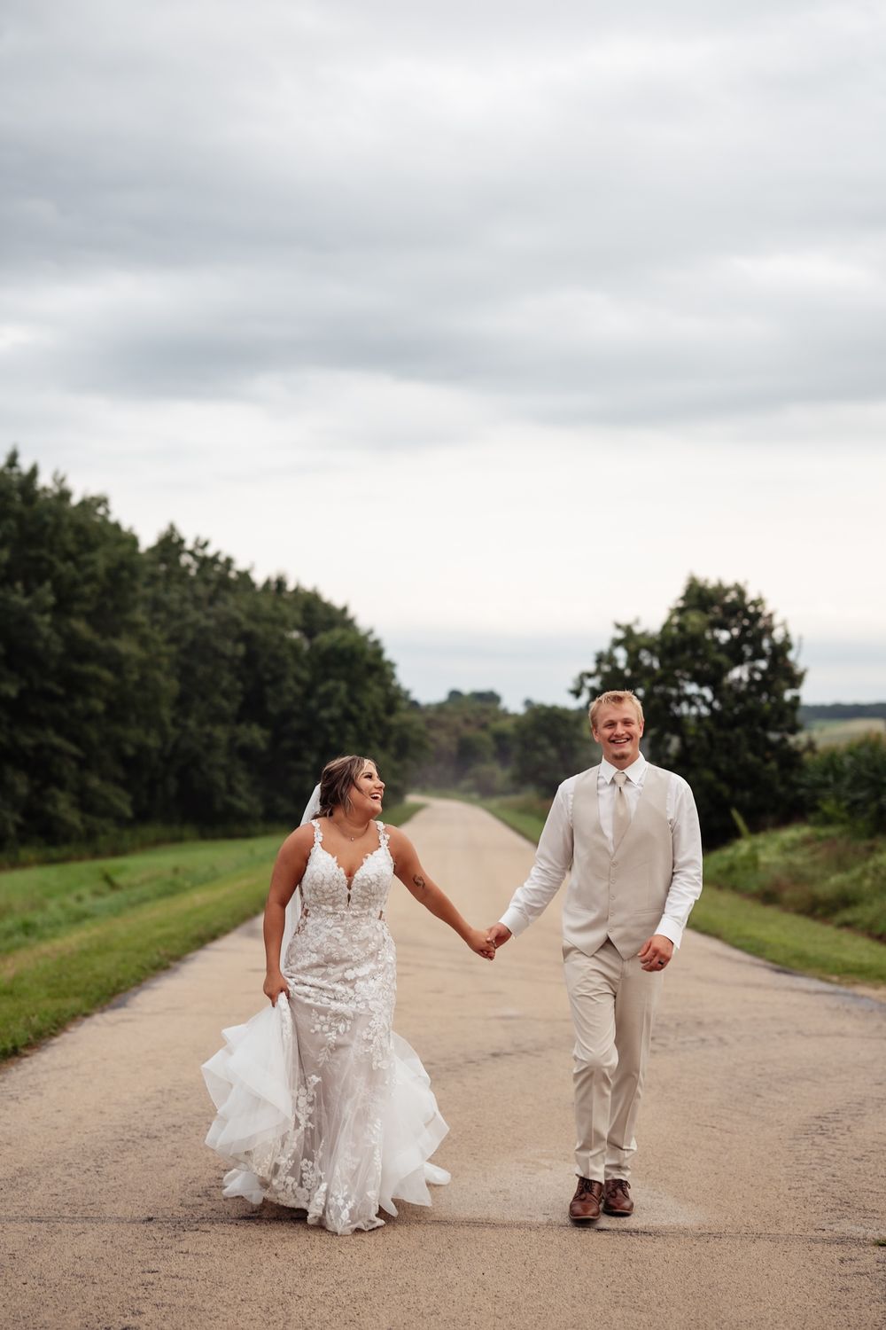 A couple in wedding attire playfully runs and laughs together on a country dirt road.