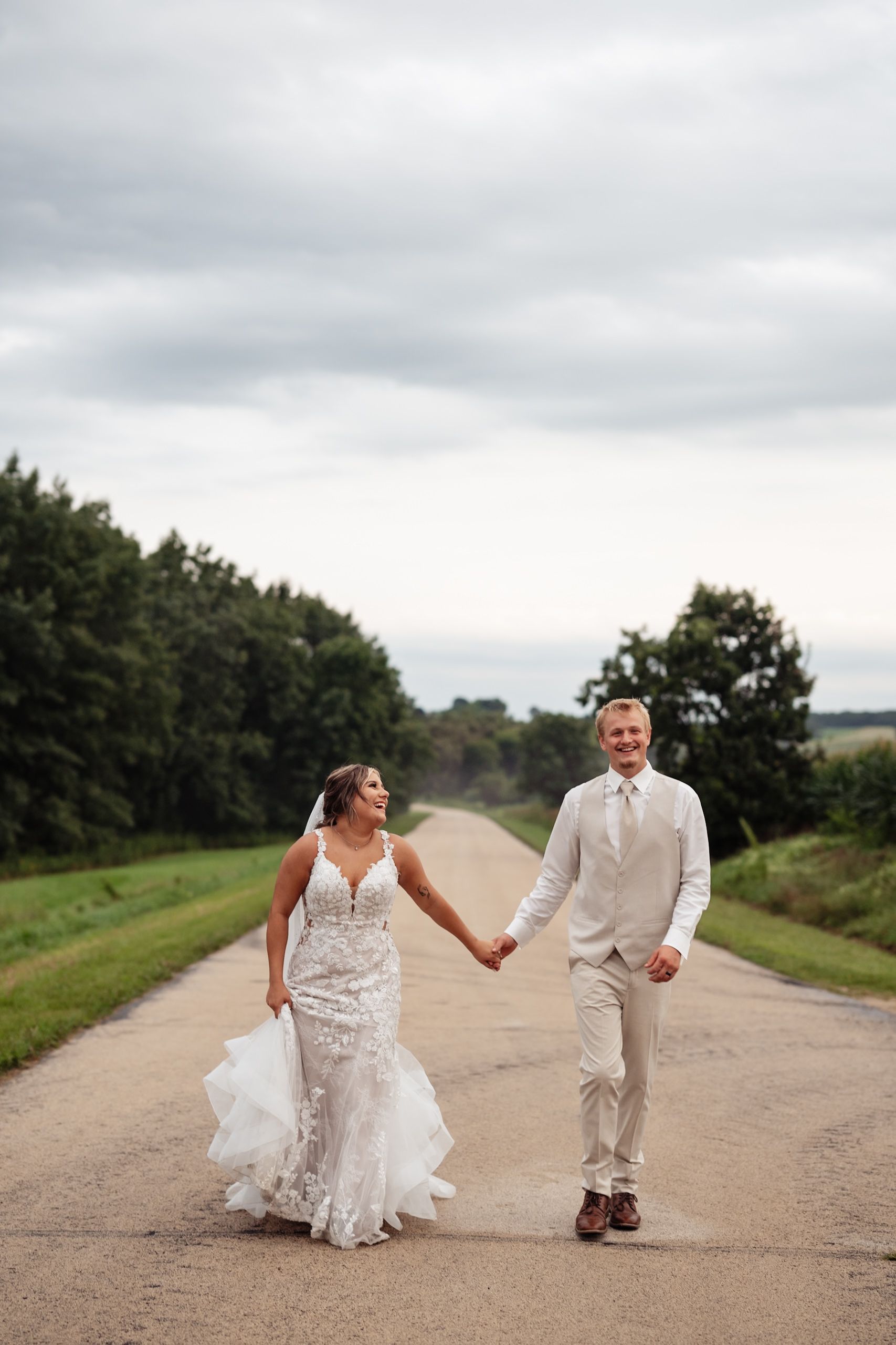 A couple in wedding attire playfully runs and laughs together on a country dirt road.
