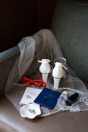 Wedding accessories including passports, rings, veil, and white shoes arranged on a grey couch.