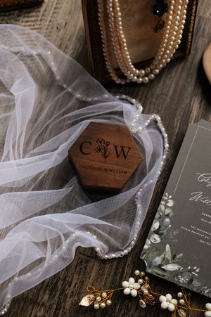 Wedding details including wooden ring box, delicate veil, floral invitation and pearl jewelry on rustic wooden surface.