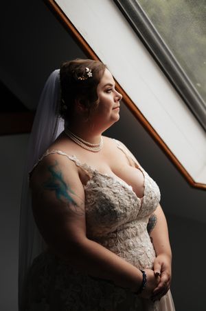 Bride in lace wedding dress with pearl necklace poses near window in dramatic natural lighting.