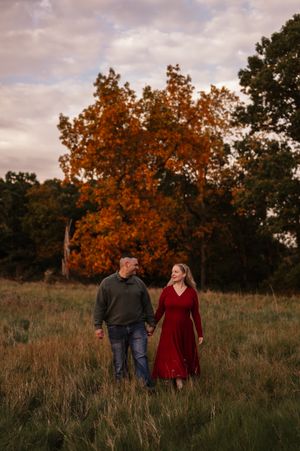 A couple walks hand in hand through a golden autumn field at sunset with tall grass and trees in the background.