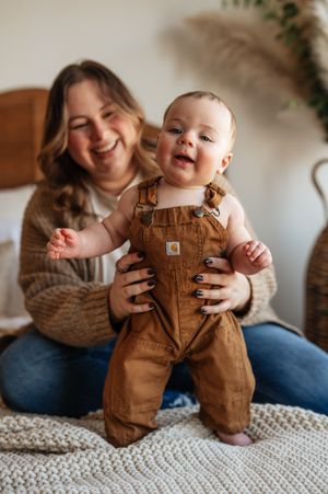 A baby wearing brown overalls sits and laughs on a textured white blanket during a lifestyle photoshoot.