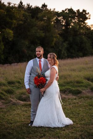 A romantic sunset wedding photoshoot in a field shows a couple in formal attire with a red bouquet.