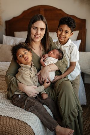 Family sits together on bed in coordinated neutral tones while holding newborn baby in intimate portrait.