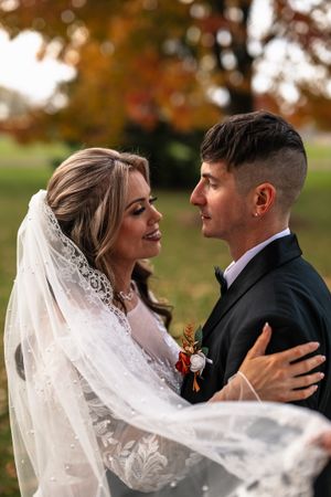 A romantic autumn wedding portrait with a bride in lace veil and groom in black suit embracing outdoors.