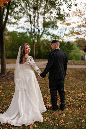 A bride and groom hold hands while standing on fallen autumn leaves in a wooded outdoor setting.