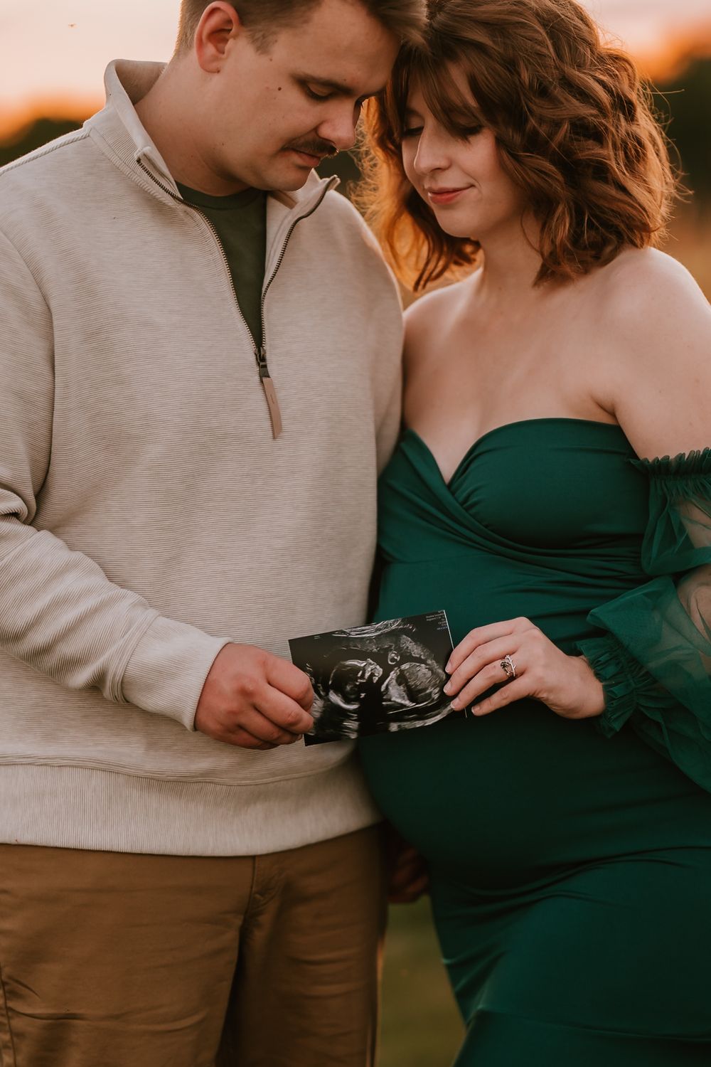 A couple in casual attire shares an intimate moment looking at an ultrasound photo during sunset.