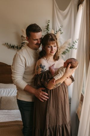 A family shares tender moments near a window with soft natural light and white curtains in a minimalist setting.