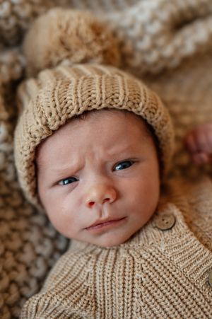 Close up portrait of a baby wearing a knitted beige hat and sweater against a textured background.