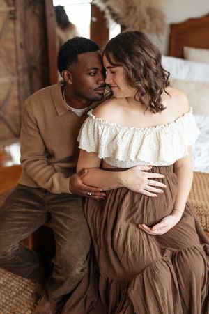 A romantic couple in brown and cream colored outfits embrace tenderly on a couch in a warm-lit indoor setting.