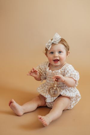 Smiling baby wearing a gingham romper and lace bow headband sits on beige backdrop.