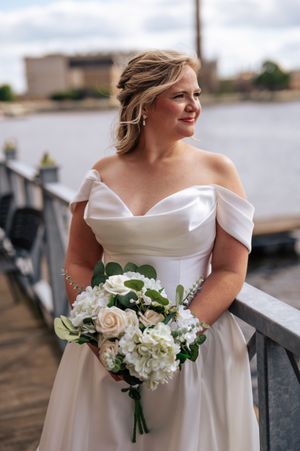 Elegant bride in off-shoulder white wedding gown holds a romantic bouquet of blush and white flowers by the waterfront.