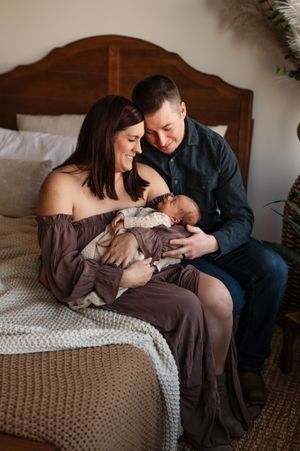 A couple sits on a bed with their newborn baby, sharing an intimate family moment in warm lighting.