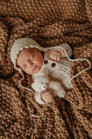 Newborn baby sleeping peacefully on brown textured blanket wearing white knit bonnet and wrap.