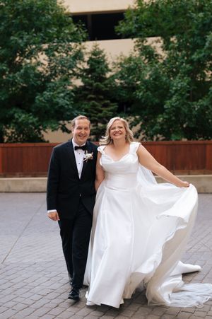 A couple in formal wedding attire walk and laugh together on a stone paved courtyard surrounded by greenery.