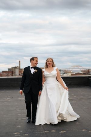 A bride and groom walk together on an urban rooftop with cloudy skies in the background.