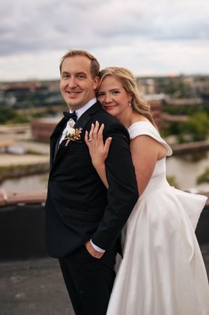A bride in a white dress shows off her ring while posing with her groom on a rooftop overlooking a cityscape.