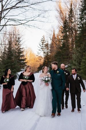 Wedding party walking together through a snowy forest path with evergreen trees and golden sunset light.