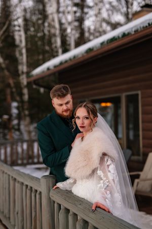 A winter wedding couple stands on a snowy cabin deck in the woods with bride wearing white fur wrap and veil.