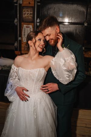 Romantic wedding couple embraces in dimly lit rustic venue with bride wearing flowing white gown with puffy sleeves.