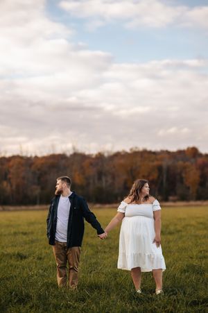 A couple in casual attire walks through a golden autumn field at sunset with fall foliage in the background.