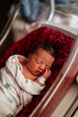 A sleeping newborn baby rests peacefully on red textured bedding in a hospital bassinet.