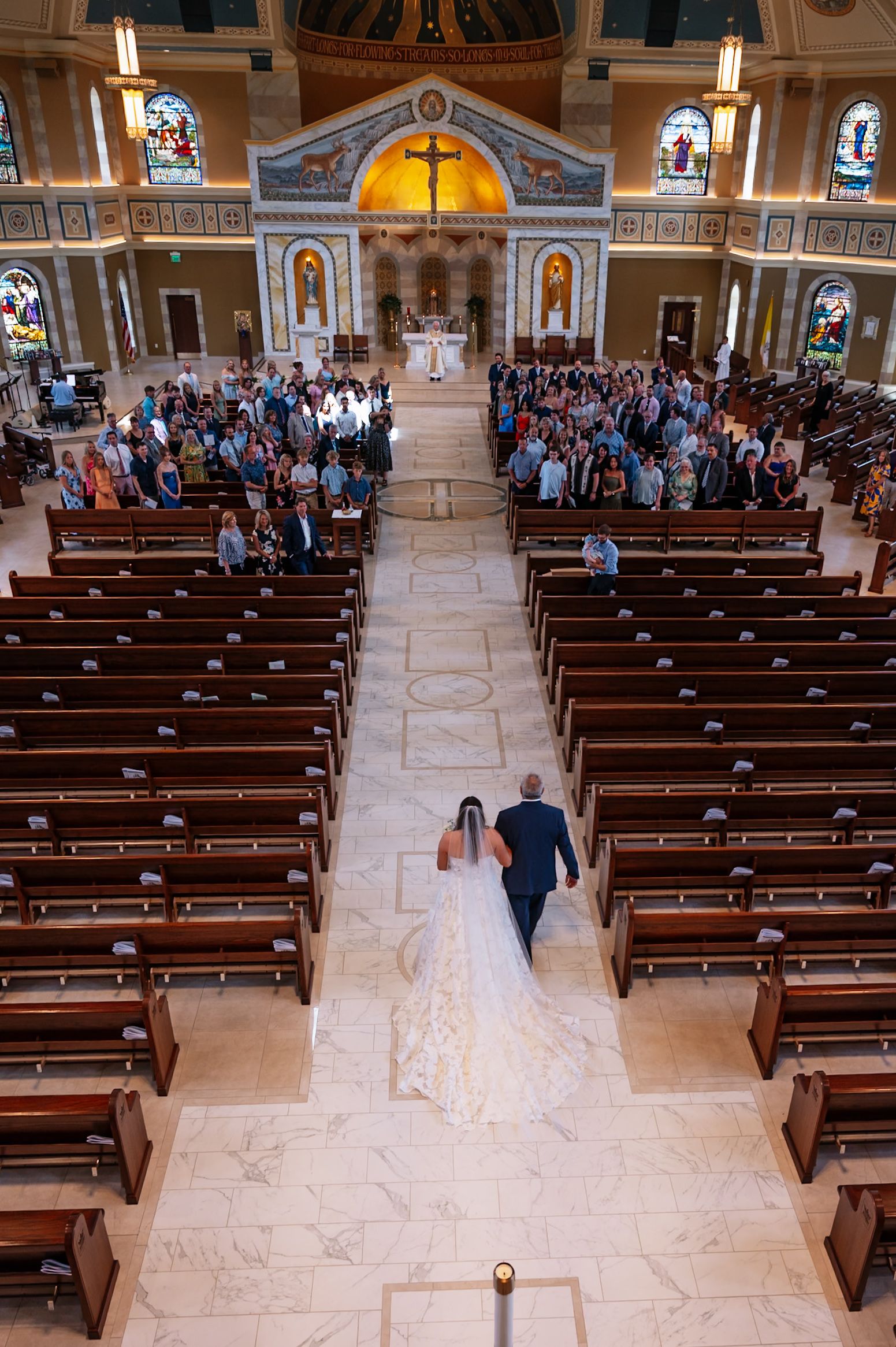 A couple walks down the center aisle of an ornate Catholic church during a wedding ceremony with guests seated in wooden pews.