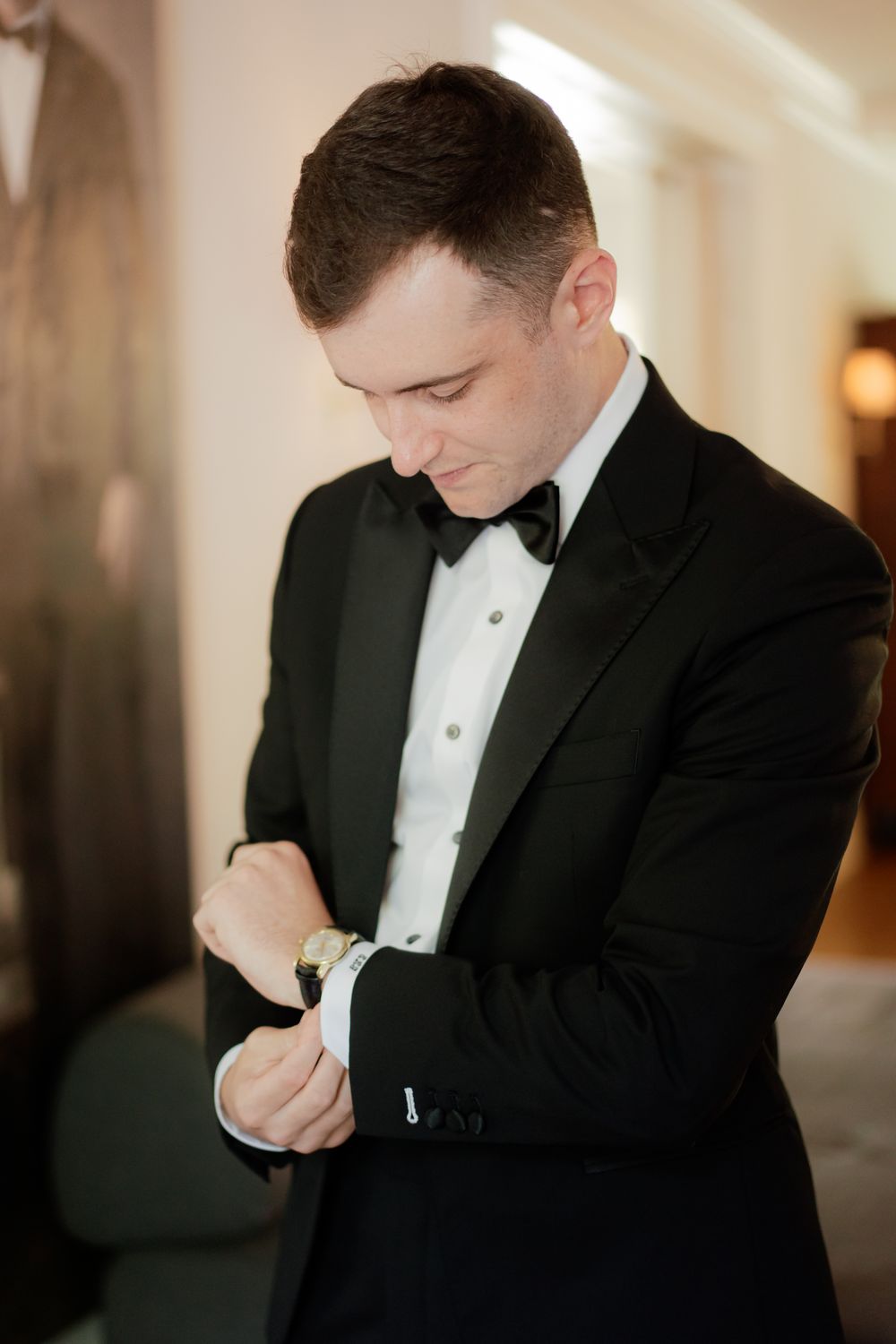 Person in formal black tuxedo checking time on wristwatch in elegant interior setting.