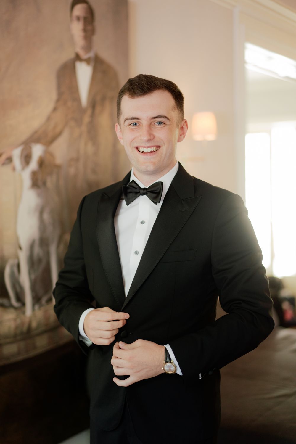 A person wearing a black tuxedo and bowtie stands smiling in front of a painting in an elegant indoor setting.