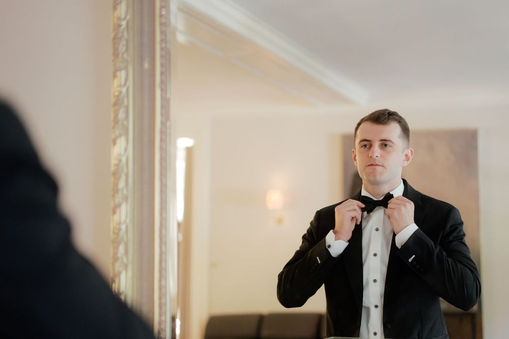 A person in a black tuxedo adjusts their bowtie in an elegant hallway with ornate gold-framed mirrors.