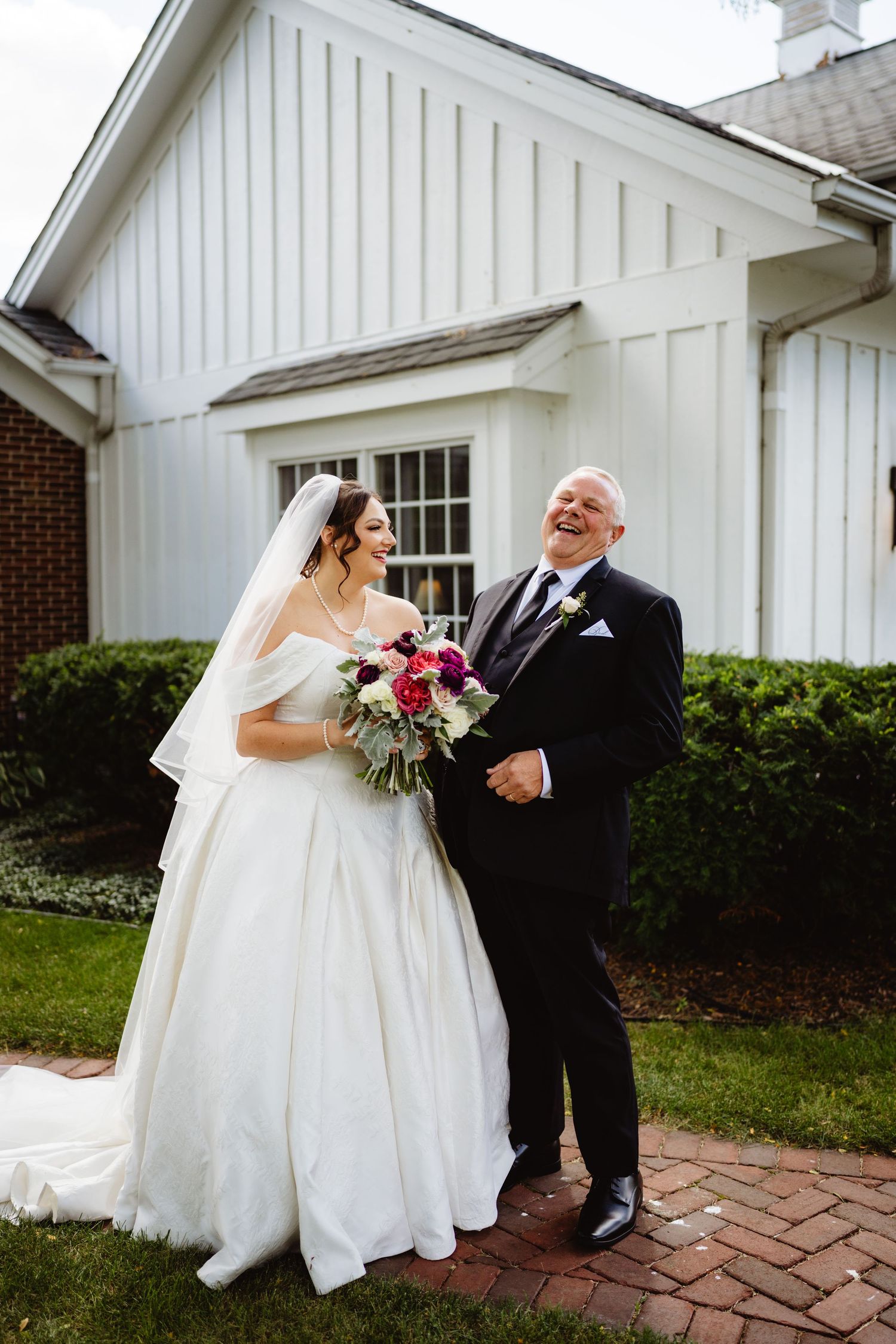 A pair in formal attire share a joyful moment together on a brick path outside a white church building.