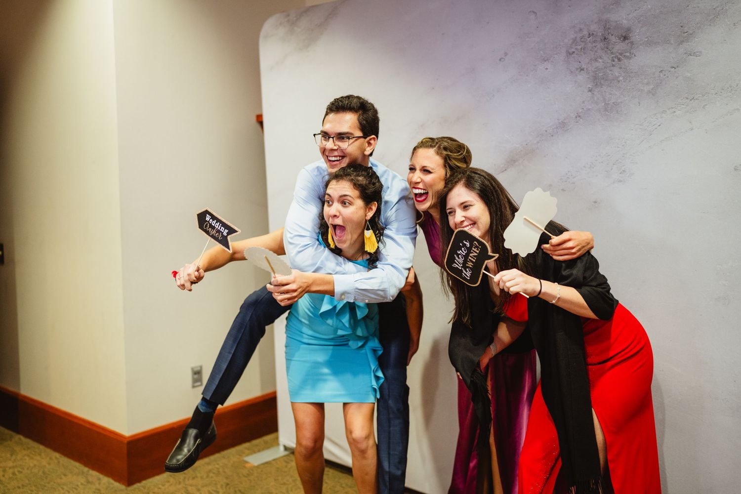 Four friends pose energetically together while holding gaming controllers and remotes in an indoor hallway.