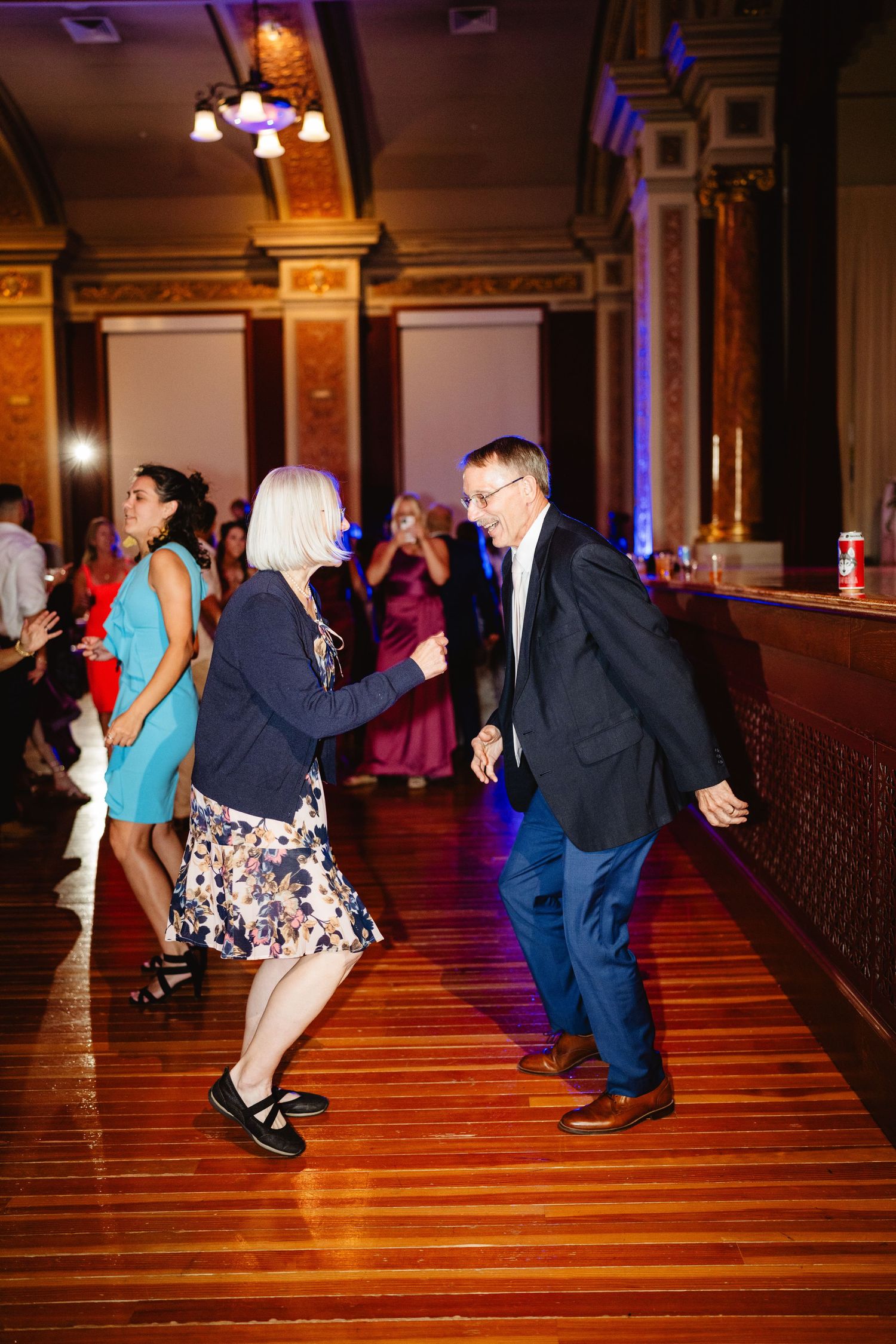 Party guests dance in a line on a polished wooden floor under ornate lighting in an elegant ballroom.