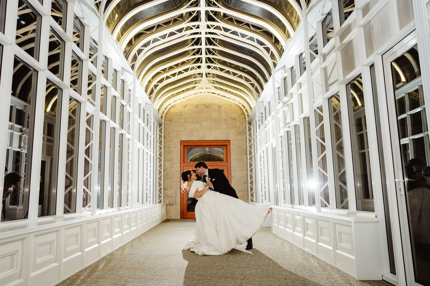 A long, white hallway with arched ceiling and elegant architectural details illuminated by warm lighting stretches into distance.